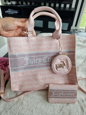 Ljusrosa handväska från Juicy Couture - Ljusrosa handväska från Juicy Couture med tryckt logotyp och matchande plånbok samt rund liten väska med JC-detalj. Väskan har både handtag och axelrem, och är tillverkad i textil med detaljer i konstläder. Perfekt för dig som gillar en söt och trendig look.