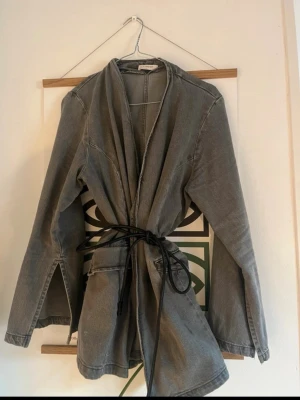 Grå jeansjacka/kavaj med knytskärp Lindex - (Slutsåld) snygg grå jeansjacka i kavajstil från Lindex med avslappnad passform och knytskärp i midjan. Jackan har öppen front utan knappar och långa ärmar med slits vid ärmslut. Perfekt för en cool och avslappnad stil.