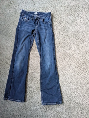 Blå bootcut jeans med fickdetaljer - Säljer ett par blå bootcut jeans med klassisk femficksdesign och snygga ficklock bak. Jeansen har ljusare slitningar framtill och kontrastsömmar. Materialet är denim i bomull. Mycket fint skick och inga synliga defekter. Storlek 164 ban motsvarar xs/s i vuxen