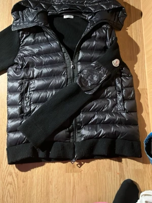 Svart dunväst från Moncler med huva - Snygg svart dunväst från Moncler med huva och stickade detaljer vid ärmhål och nederkant. Västen har dragkedja framtill, två fickor med knapp  och klassisk Moncler-logga på vänster arm.Perfekt för lager-på-lager och streetstyle. Saknar dock knapparna på båda fickorna Det är bara att fråga om du undrar nåt pris kan diskuteras vid snabbt köp 