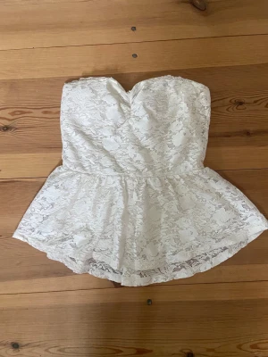 Vit spets tubtopp - Säljer en söt vit axelbandslös tubtopp med peplumskärning och spetsdetaljer. Toppen har en hjärtformad urringning och är fodrad under spetsen för extra komfort. Perfekt för dig som vill ha en romantisk och trendig look.