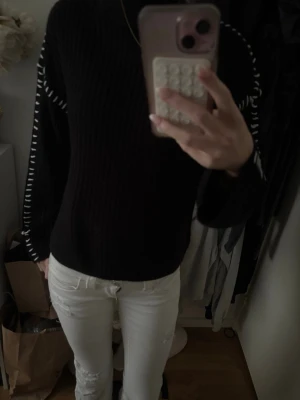 Svart stickad tröja med vita detaljer - Svart stickad tröja med ribbad struktur och vita dekorativa sömmar längs ärmarna. Tröjan har hög hals och en avslappnad passform, perfekt för en cool och enkel look. 