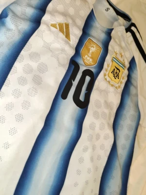 Argentina matchtröja Adidas nr 10 - Säljer en officiell Argentina fotbollströja från Adidas med klassiska blå och vita ränder, guldiga detaljer och nummer 10 på bröstet. Tröjan har FIFA 2022-märke och AFA-emblem med tre stjärnor. Perfekt för fotbollsfans!