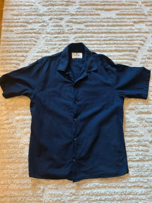 Mörkblå skjorta från Skagen - Stilren mörkblå skjorta från Skagen med korta ärmar och klassisk krage. Skjortan har knappar framtill och är tillverkad i ett mjukt linnematerial som passar perfekt till sommaren. Enkel och clean design som funkar till många olika looks.