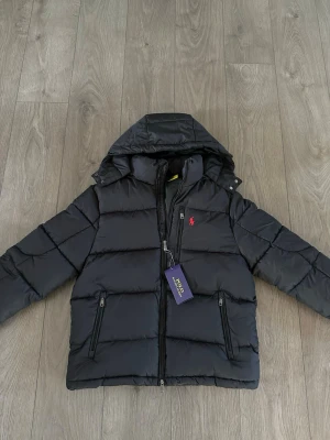 Ralph Lauren svart - Svart pufferjacka från Polo Ralph Lauren med huva, två dragkedjefickor och röd logga på bröstet. Jackan har quiltad design och dragkedja framtill. Perfekt för kalla dagar och riktigt snygg streetstil.