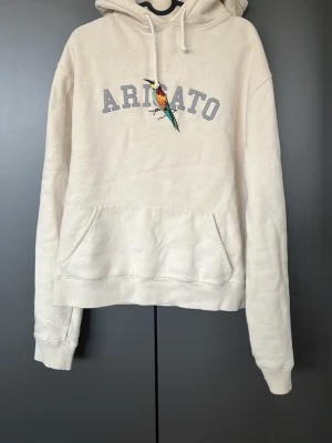 Beige hoodie från Axel Arigato - Snygg beige hoodie från Axel Arigato med broderad fågel och texten 'ARIGATO' på bröstet. Hoodien har huva med dragsko och en stor magficka. Perfekt för en chill och stilren look. Tillverkad i mjukt material för extra komfort.