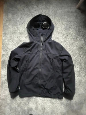 Svart vindjacka från C.P. Company - Snygg svart vindjacka från C.P. Company med ikoniska glasögon i huvan. Jackan har dragkedja framtill, två sidofickor med dragkedja och justerbar nederkant. Tillverkad i ett lätt och slitstarkt material, perfekt för blåsiga dagar.