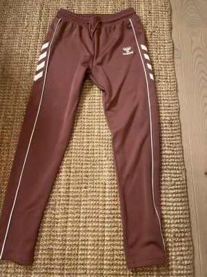 Bruna mjukisbyxor från Hummel - Säljer ett par bruna mjukisbyxor från Hummel med vita detaljer och logga på vänster lår. Byxorna har vita ränder längs benen och elastisk midja med dragsko. Tillverkade i ett mjukt och stretchigt material som passar perfekt för chill eller träning. Men det är en tråd som har gått upp på rumpan, eller hur man ska förklara det, jag har med en bild också.