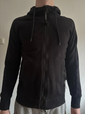 Svart hoodie med dragkedja från C.P. Company - Säljer en svart hoodie från C.P. Company med dragkedja framtill och två fickor med dragkedja. Huvan har justerbara snören och är utrustad med de ikoniska glasögon-linserna baktill. OPS storlek L men passar som en M