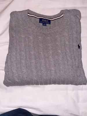 Grå kabelstickad tröja Polo Ralph Lauren - Snygg grå kabelstickad tröja från Polo Ralph Lauren med rund halsringning och klassisk broderad logga på bröstet. Tröjan passar till alla tillfällen i vardagen och är även i väldigt bra skick. Ny pris är 1599 kr! Hör av dig vid funderingar!