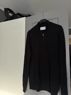 Svart halvzip tröja Selected Homme - En svart långärmad tröja från Selected Homme med halvzip och hög krage. Tröjan är enkel och stilren, perfekt för att matcha med jeans eller chinos. Materialet är mjukt och bekvämt, och designen är klassisk med ribbade muddar.