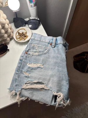 Ljusblåa slitna jeansshorts - Säljer ett par ljusblåa jeansshorts med råa, fransiga kanter och flera slitningar framtill. Klassisk femficksmodell med knappgylf. Perfekta för sommaren och ger en avslappnad vibe.🤍