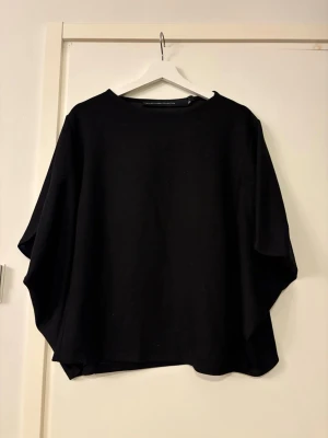 Svart loose topp prislapp kvar  - En svart, loose-fit topp med breda, fladdriga ärmar och rund halsringning. Toppen har en minimalistisk design och är tillverkad i ett mjukt material som känns skönt mot huden. Perfekt för en avslappnad och stilren look. Prislapp kvar strl M