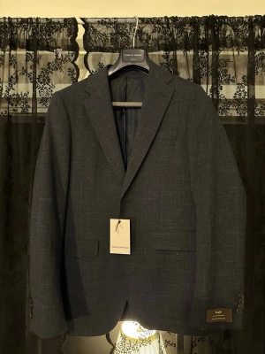 Mörkblå kavaj från These Glory Days - Nypris 3999 kr. HELT OANVÄND! Stilren mörkblå kavaj från These Glory Days med diskret rutmönster. Klassisk passform med två framfickor och knappar vid ärmslut. Tillverkad i ett tunt, vävt material som ger en modern look. Perfekt för dtudent eller bal! 