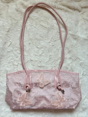 Rosa handväska med spets och fjärilar - Söt rosa handväska med lång axelrem, dekorerad med spets och broderade fjärilar på framsidan. Väskan har en rektangulär form och detaljer i matchande rosa nyans. Perfekt för dig som gillar gulliga accessoarer och vill sticka ut lite extra.
