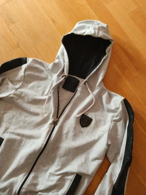 Grå hoodie + byxa  - Snygg grå hoodie från Philip plane med svarta ribbade detaljer längs ärmarna och svarta muddar. Dragkedja framtill, två fickor och en svart badge på bröstet. Huvan har vita snören och svart innerfoder. Perfekt för en streetwear-look.på byxan märket är lite sönder och vid midjan men lagat de på midjan du ser bilderna 