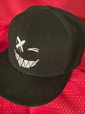 Svart keps med broderad smiley - Säljer en svart keps med platt skärm och ett broderat vitt smiley-ansikte framtill. Designen har ett stort leende med spetsiga tänder och ett kryssat öga, vilket ger en edgy och lekfull look. Kepsen är tillverkad i bomullstyg och har klassisk snapback-form.