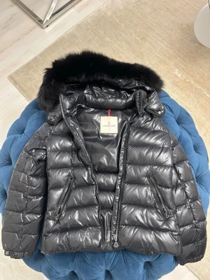 Svart dunjacka med pälskrage Moncler - Snygg svart dunjacka från Moncler med glansig finish och avtagbar huva med svart pälskant. Jackan har två snedställda dragkedjefickor fram och quiltad design. Perfekt för kalla vinterdagar och riktigt trendig look.