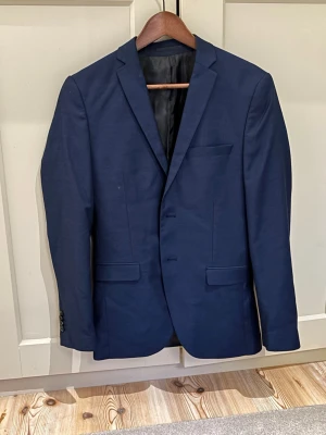 Mörkblå kostym med kavaj och byxor - Snygg mörkblå kostym med klassisk kavaj och matchande byxor. Kavajen har två knappar, smala slag och fickor med lock. Byxorna har raka ben och bälteshällor. Perfekt för dig som vill ha en stilren och tidlös look. Strlk 44 i kavaj. Strlk 46 i byxor.