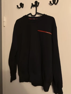 Svart sweatshirt från Prada - Svart sweatshirt från Prada med rund hals och lång ärm. Diskret röd detalj med logotyp på bröstet. Klassisk och enkel design som passar perfekt till jeans eller joggers.