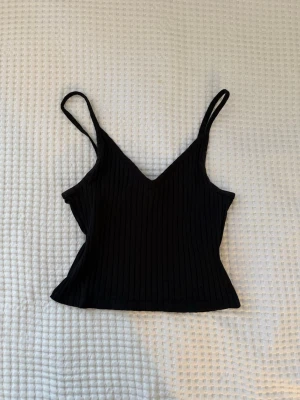 Svart ribbad linnetopp - Säljer en svart ribbad linnetopp med smala axelband och v-ringad hals. Toppen är croppad och har en tight passform, perfekt till jeans eller kjol. Enkel och stilren design som funkar till många olika looks.