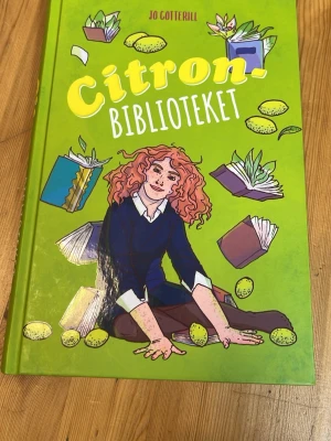 Citronbiblioteket av Jo Cotterill - Säljer boken 'Citronbiblioteket' av Jo Cotterill. Boken har ett hårt grönt omslag med illustrationer av böcker, citroner och en tjej med lockigt rött hår. Perfekt för dig som gillar ungdomslitteratur och berättelser om vänskap och familj.