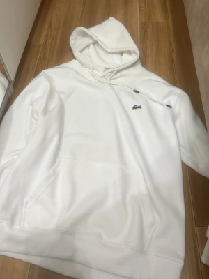 Vit hoodie från Lacoste med huva - Säljer en stilren vit hoodie från Lacoste med klassisk krokodillogga på bröstet. Hoodien har huva med dragsko och en stor magficka framtill. Perfekt för dig som gillar enkel och clean streetwear.