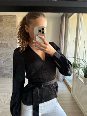 Svart omlottblus med knytband - Snygg svart omlottblus med långa ärmar och markerade axlar. Blusen har ett knytband i midjan som ger en fin siluett! Använd 2 gånger, sitter som en xs 💕