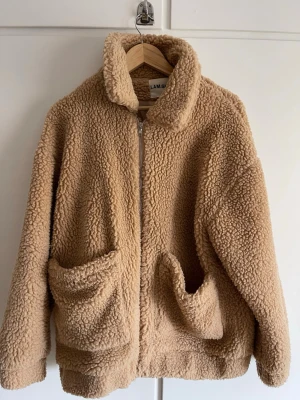 Beige teddyjacka från I.AM.GIA - Mysig beige teddyjacka från I.AM.GIA med dragkedja framtill och två stora fickor. Jackan har hög krage och är gjord i fluffigt teddy-material som håller dig varm och trendig. Perfekt för kyliga dagar och enkel att matcha med olika outfits. -oversize