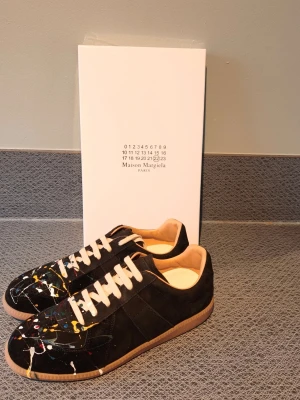 Maison Margiela svart sneakers grafitti - Maison Margiela sneakers i svart mocka och läder med unika färgstänk i vitt, gult och rött. Klassisk låg modell med vita snören och beige sula. Snygg och konstnärlig design som sticker ut. Kommer med originalkartong.