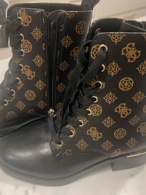 Svarta stövletter från Guess - Snygga svarta stövletter från Guess med guldfärgade logomönster över hela skon. Boots i konstläder med snörning framtill, dragkedja på sidan och gulddetaljer vid hälen. Perfekta soppar för dig som vill sticka ut.