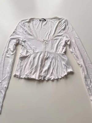 Vit långärmad topp med spetsdetaljer - Söt vit långärmad topp från ginatricot med spetskant runt ringningen och rosett framtill. Toppen har en liten rosett vid urringningen och volangkant nedtill. Materialet känns mjukt och lätt, perfekt för en trendig och feminin look.