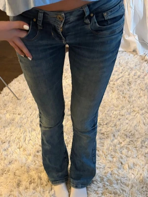 Blå bootcut jeans med låg midja - Säljer ett par LTB blå bootcut jeans med låg midja och klassiska fem fickor. Jeansen har en snygg tvättad look och stängs med knapp och dragkedja. Perfekta för en avslappnad och trendig stil. Defekt på ena bakfickan (inget man tänker på)