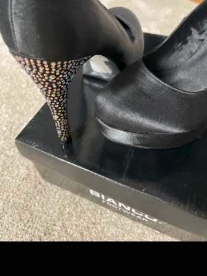 Svarta pumps från Bianco med strass - Snygga svarta pumps från Bianco i satinliknande material. Klacken är täckt med glittrande strass i olika färger som ger extra wow-effekt. Klassisk rund tå och elegant siluett, perfekta för att sticka ut på nästa event.