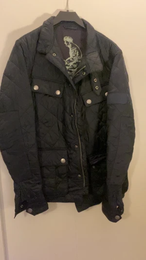 Svart quiltad dunjacka med tryckknappar - Säljer en svart quiltad dunjacka med coola tryckknappar och dragkedja framtill. Jackan har två bröstfickor och två sidofickor, samt detaljer vid ärmsluten. Perfekt för kalla vinterdagar och riktigt snygg till streetwear-stilen.