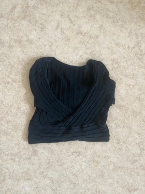 Svart omlottopp med ribbad struktur - Snygg svart omlottopp med ribbad struktur och långa ärmar. Toppen är croppad och har en djup v-ringning framtill. Perfekt att styla med  jeans eller kjol.