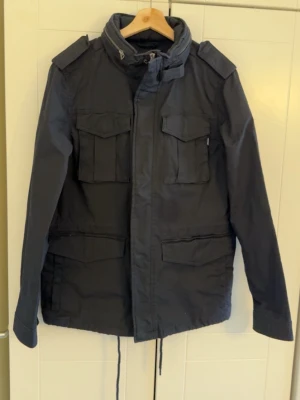 Svart field jacket  - Stilren svart field jacket med fyra stora fickor framtill, dragsko i midjan och hög krage. Jackan har axelklaffar och stängs med både dragkedja och tryckknappar. Perfekt för allround dagar och snygg till streetwear. Det finns snören vid midjan som gör så att den inte sitter för löst men man kan och ta bort de om de skulle vara till besvär