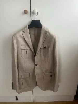 Beige kavaj från The Tailoring Club - Stilren beige kavaj från The Tailoring Club i storlek 46 en ruskigt snygg safariblazer. Tillverkad i ett grovt vävt material med fyra fickor framtill och mörka knappar. Klassisk krage och enkelknäppt design som ger en avslappnad men ändå dressad look. Nypris 3500 kr 