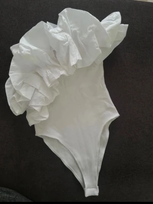 Eftertraktad Vit body med volang från Zara - Supersnygg vit body från Zara med stor volang över ena axeln. Tight passform och mjukt material i bomullsblandning. Perfekt för dig som vill sticka ut med en unik och trendig look.