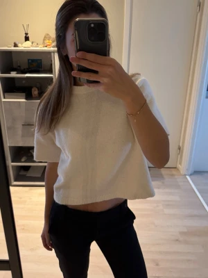 Vit/beige croppad stickad topp - Säljer en vit/beige croppad stickad topp med korta ärmar och rund hals. Perfekt för dig som gillar en enkel och clean stil. Toppens stickade material ger en mjuk och skön känsla och den är lätt att matcha med olika outfits. Säljer då den inte kommer till användning. Använd ett fåtal gånger.