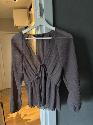 Plisserad blus från Zara - Trendig plisserad blus i lila från Zara med djup v-ringning och knytning framtill. Blusen har långa ärmar och är tillverkad i ett lätt, transparent material som ger en luftig känsla. Perfekt att styla med jeans eller kjol för en cool look.