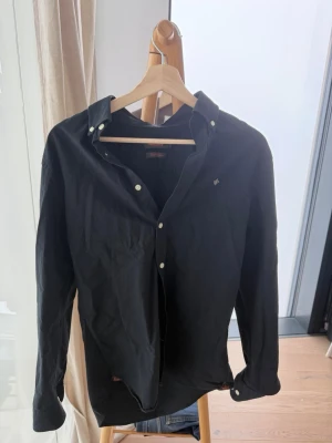 Svart skjorta med knappar - Klassisk svart skjorta med långa ärmar och vita knappar. Skjortan har button-down krage och en liten broderad detalj på bröstet. Perfekt till jeans eller chinos för en stilren look.