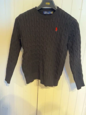 Svart kabelstickad tröja Polo Ralph Lauren - Snygg svart kabelstickad tröja från Polo Ralph Lauren med klassisk ranges röd logga broderad på bröstet. null har rund hals double ribbade muddar vid ärmslut och nederkant. Perfekt för dig som gillar tidlös och stilren design.