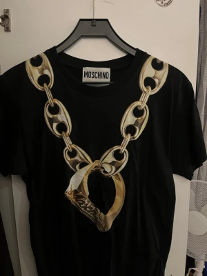 Svart Moschino t-shirt med kedjetryck - Säljer en svart t-shirt från Moschino med ett stort tryck av en guldfärgad kedja och ring framtill. T-shirten har rund hals och korta ärmar. Perfekt för dig som vill sticka ut med en unik och trendig design.