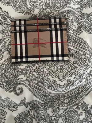 Burberry rutigt kortfodral i beige - Snyggt kortfodral från Burberry i klassiskt beige med svart, vitt och rött rutmönster. Tillverkat i slätt läder med flera kortfack och diskret Burberry-logga framtill. Perfekt för dig som vill ha en stilren och ikonisk accessoar.