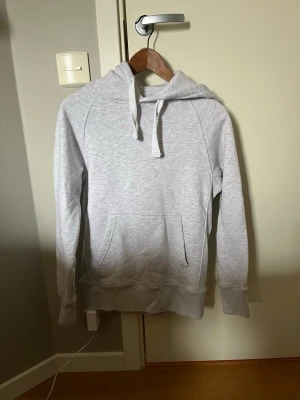 Russemerch  - En stilren ljusgrå hoodie med huva och dragsnören. Hoodien har en stor magficka och ribbade muddar vid ärmslut och nederkant. Perfekt för en avslappnad och casual look. Materialet känns mjukt och bekvämt, troligen bomullsblandning.