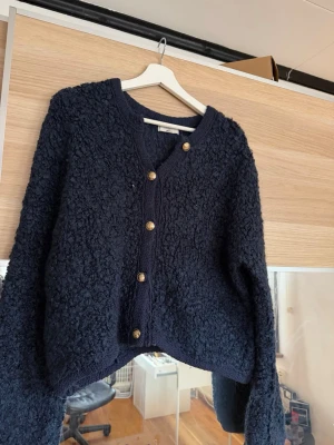 Mörkblå teddykofta från Gina Tricot - Mysig mörkblå teddykofta från Gina Tricot med dekorativa guldfärgade knappar framtill. Koftan har v-ringning, lång ärm och en fluffig, strukturerad yta som ger en trendig look. Perfekt för lager på lager och att hålla värmen under kyliga dagar.