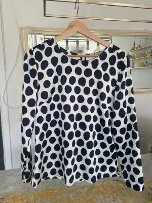 Vit blus med stora svarta prickar - Snygg vit blus med stora mörkblå prickar och rund halsringning. Blusen har långa ärmar och knäppning med tygklädda knappar i ryggen. Tillverkad i ett mjukt, lätt material som ger en skön känsla. Perfekt för dig som gillar ett lekfullt och trendigt mönster.