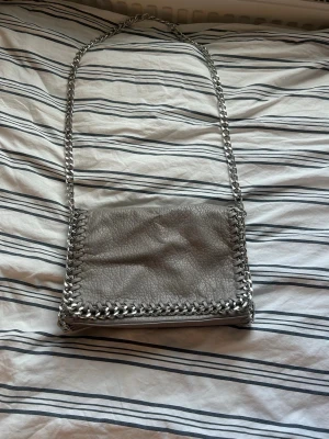 Grå axelväska från Tiamo - Snygg grå axelväska från Tiamo.  Har en  silverkedja som axelrem och runt kanterna.  Väldigt bra i skick och knappast använd. 