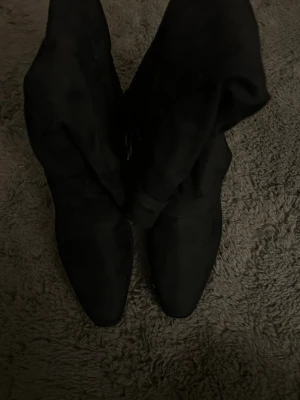 Svarta mockastövlar med silverdetalj - Snygga svarta stövlar i mocka med spetsig tå och dekorativa silverfärgade detaljer upptill. Stövlarna har en klassisk och stilren design som passar till många outfits. Perfekta för dig som vill ha ett par trendiga boots med det lilla extra.